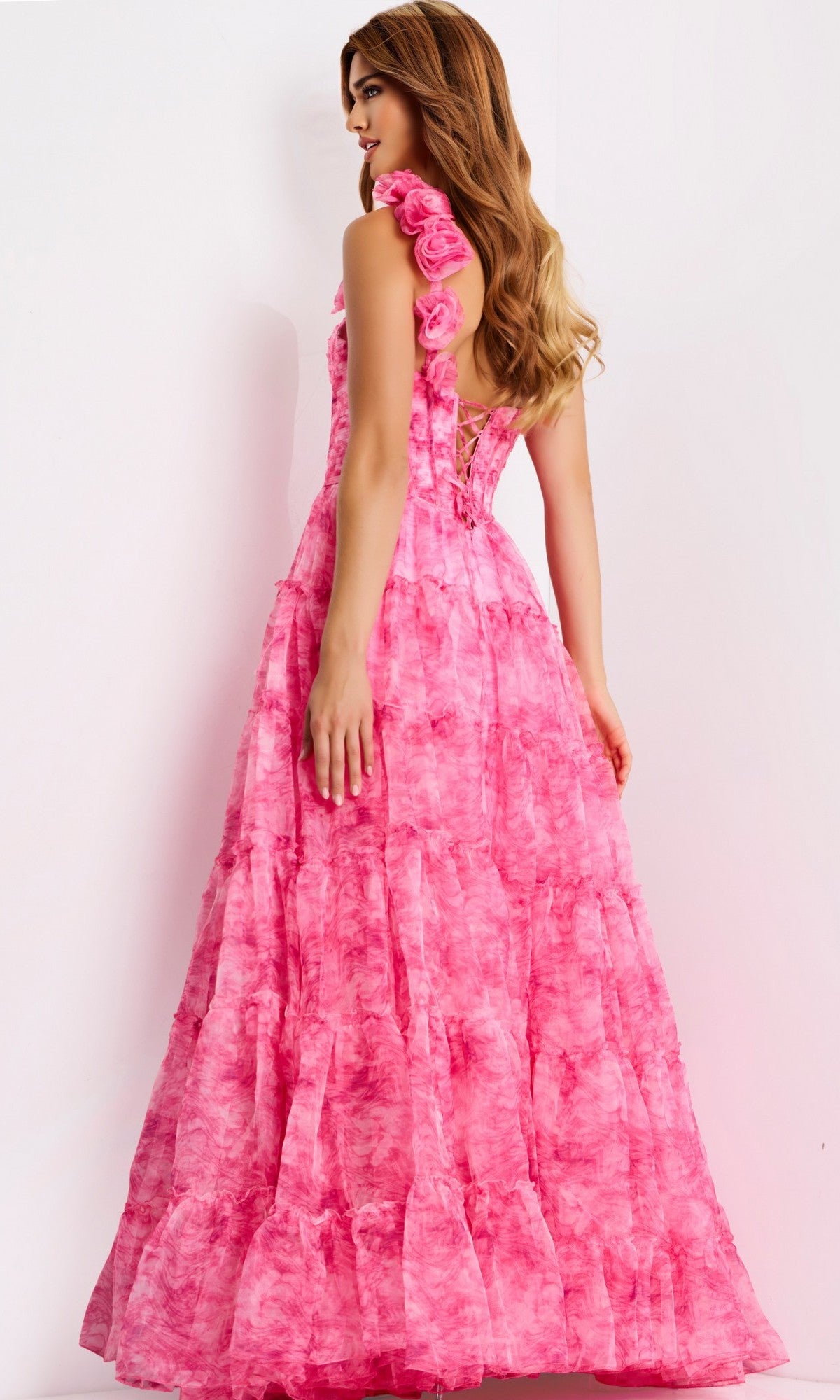 A woman wears the Jovani 49074 long prom dress, a sleeveless, floor-length pink gown with layered fabric, rose shoulder details, and a subtle corset bodice, standing against a plain light background.